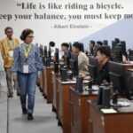 Two instructors walk through a computer lab as students work at desks with an einstein quote on the wall life is like riding a bicycle